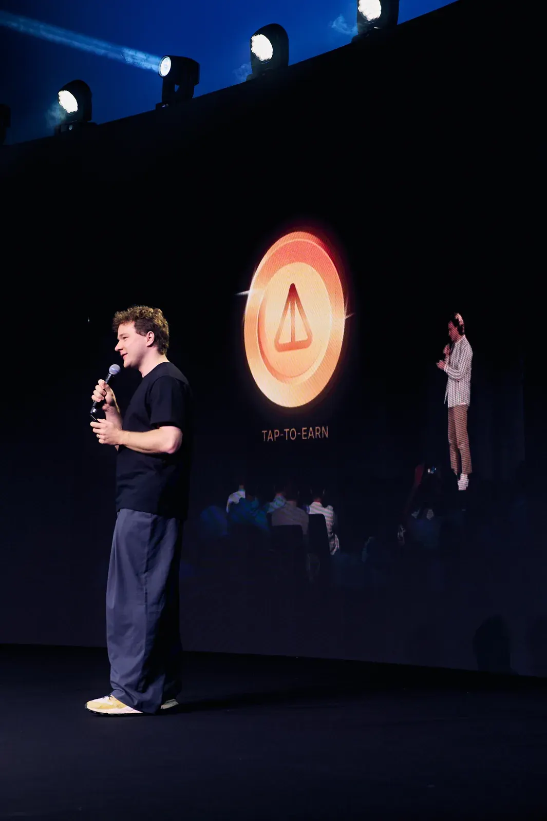 Speaker with microphone on stage at a Dubai investment event, with audience and TAP-TO-EARN screen behind Speaker with microphone on stage at a Dubai investment event, with audience and TAP-TO-EARN screen behind